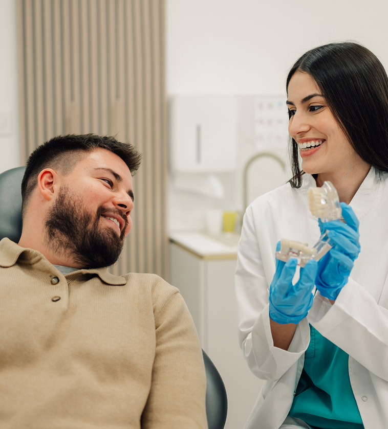 A smiling female dentist in a white lab coat and blue gloves holds a clear anatomical model of teeth while talking to a seated male patient. Both are smiling in a bright, modern dental office.